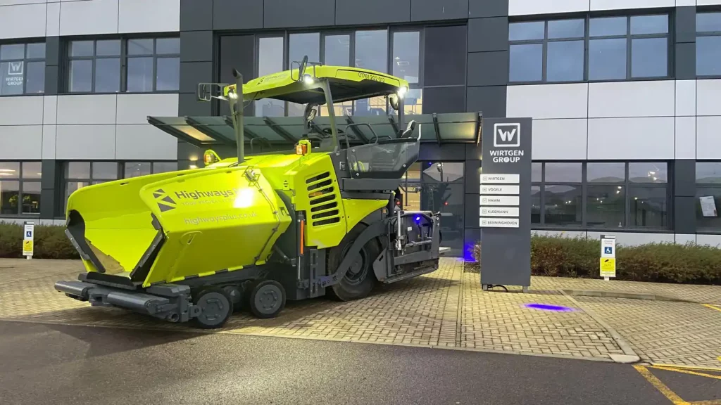 A bright yellow road paver machine, ideal for motorways and faster project completion, is parked on a paved area outside a modern office building with large windows and a sign that reads "Wirtgen Group. | Highways Plus